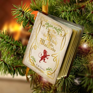 May include: A miniature book ornament titled The Lion, the Witch & the Wardrobe by C.S. Lewis, featuring a cream-colored cover with gold filigree, a red lion illustration, and a gold ribbon loop for hanging on a Christmas tree branch.