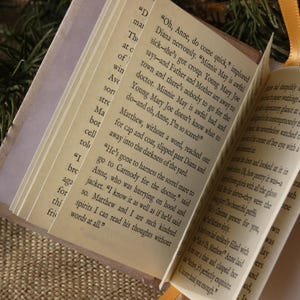 May include: A miniature book with cream-colored pages featuring printed black text from the novel Anne of Green Gables. The small volume has a light-colored cover and a thin orange ribbon bookmark attached to the top edge of the spine.