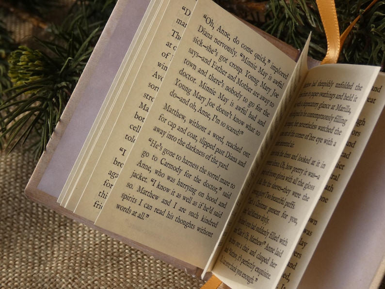 May include: A miniature book with cream-colored pages featuring printed black text from the novel Anne of Green Gables. The small volume has a light-colored cover and a thin orange ribbon bookmark attached to the top edge of the spine.