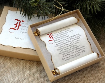 May include: A scroll ornament featuring the text of Isaiah 9:6-7 printed in black with a red decorative initial letter. The scroll has burnt edges, gold-toned end caps, and a gold hanging cord, presented inside a square cardboard gift box.