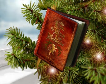 May include: A miniature book ornament titled The Lion, the Witch & the Wardrobe by C.S. Lewis, featuring a textured reddish-brown cover with a gold lion illustration and green leaf accents, suspended by a gold ribbon from a pine tree branch.
