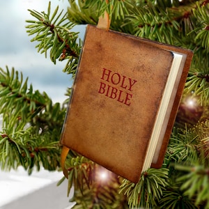 May include: A miniature Holy Bible ornament with a distressed brown leather-like cover and red embossed text, featuring a small gold ribbon loop for hanging, suspended within the green needles of a pine tree branch.