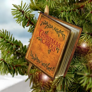 May include: A rectangular Christmas ornament shaped like a small book with a tan cover, dark brown edges, and green leaf illustrations. The text "CHRISTMAS CAROLS" is printed in red on the front cover. A gold ribbon is attached to the top.