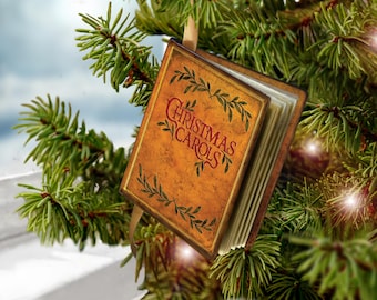 Peut inclure: Une décoration de Noël rectangulaire en forme de petit livre avec une couverture beige, des bords marron foncé et des illustrations de feuilles vertes. Le texte "CHRISTMAS CAROLS" est imprimé en rouge sur la couverture. Un ruban doré est fixé.