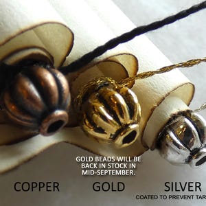 May include: Three ribbed beads in copper, gold, and silver tones, each attached to a light-colored scroll. Text below reads: COPPER, GOLD, SILVER, COATED TO PREVENT TARNISHING, and GOLD BEADS WILL BE BACK IN STOCK IN MID-SEPTEMBER.