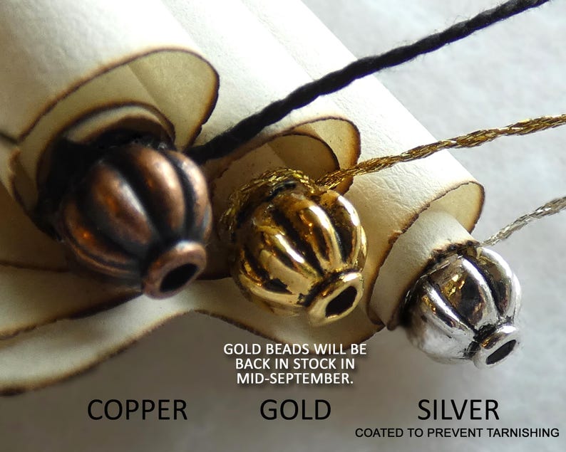May include: Three ribbed beads in copper, gold, and silver tones, each attached to a light-colored scroll. Text below reads: COPPER, GOLD, SILVER, COATED TO PREVENT TARNISHING, and GOLD BEADS WILL BE BACK IN STOCK IN MID-SEPTEMBER.