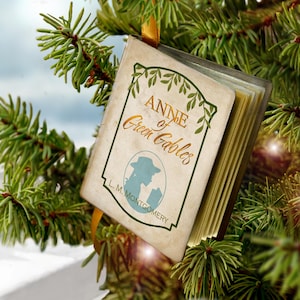 May include: A miniature book ornament titled Anne of Green Gables by L. M. Montgomery, featuring a cream-colored cover with green leaf illustrations, a silhouette of a person in a hat, and gold-toned pages, suspended by a yellow ribbon.