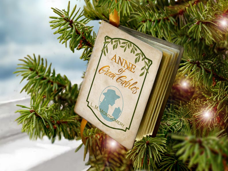 May include: A miniature book ornament titled Anne of Green Gables by L. M. Montgomery, featuring a cream-colored cover with green leaf illustrations, a silhouette of a person in a hat, and gold-toned pages, suspended by a yellow ribbon.