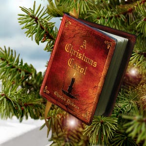 May include: A miniature book ornament titled A Christmas Carol by Charles Dickens, featuring a red cover with a black candle illustration, gold lettering, and a gold ribbon hanger, suspended among green pine needles.