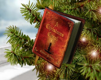 May include: A miniature book ornament titled A Christmas Carol by Charles Dickens, featuring a red cover with a black candle illustration, gold lettering, and a gold ribbon hanger, suspended among green pine needles.