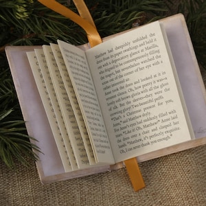 May include: A miniature book with a light-colored cover and a yellow ribbon bookmark. The open pages display printed English text, including the sentence: Matthew had sheepishly unfolded the dress from its paper swathings and held it.