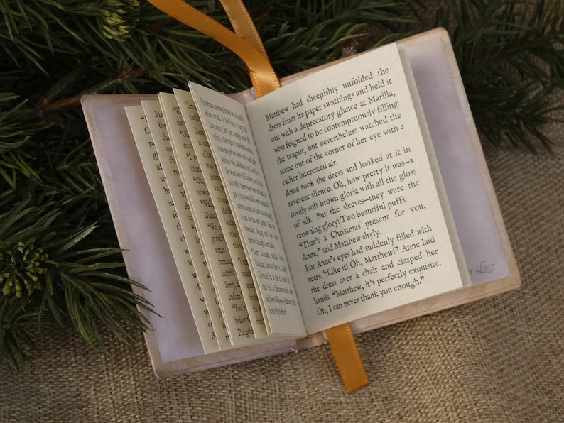 May include: A miniature book with a light-colored cover and a yellow ribbon bookmark. The open pages display printed English text, including the sentence: Matthew had sheepishly unfolded the dress from its paper swathings and held it.