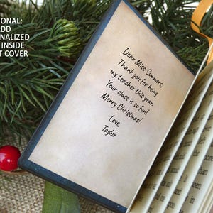 May include: A small, open book with a dark cover and cream-colored pages. The front page features printed text that reads: Dear Miss Summers, Thank you for being my teacher this year. Your class is so fun! Merry Christmas! Love, Taylor.