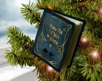 May include: A dark blue, book-shaped Christmas ornament featuring gold filigree borders and the text "Unto Us a Child is Born" in gold lettering. Below the text is a silhouette of three figures on camels under a bright star.