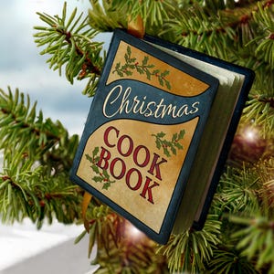 May include: A miniature book-shaped Christmas ornament hanging from a pine branch. The cover is dark blue and tan with the text "Christmas COOK BOOK" in red and white lettering, decorated with green holly leaves and red berries.
