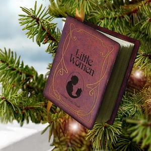 May include: A miniature book ornament titled Little Women by Louisa May Alcott, featuring a deep red cover with gold filigree accents and a black silhouette illustration of a person reading, suspended by a gold ribbon.