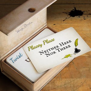 May include: A rectangular box with a hinged lid containing a stack of white cards. The top card features the green text Placey Place, black text Neither Here Nor There, a black inkwell with a yellow quill, and a green ink splatter graphic.