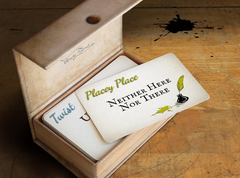 May include: A rectangular box with a hinged lid containing a stack of white cards. The top card features the green text Placey Place, black text Neither Here Nor There, a black inkwell with a yellow quill, and a green ink splatter graphic.