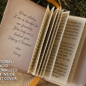 May include: A miniature book with off-white pages featuring printed text. The inside front cover displays the message: Dear Audrey, I am so thankful for our friendship. You truly are a kindred spirit. Merry Christmas! Love, Penny. The book has a yellow ribbon.