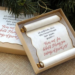 May include: A Christmas ornament in a box, featuring a scroll with text in Greek and English: "Fear not, for behold I bring you tidings of great joy!" The scroll is rolled at both ends with gold accents.