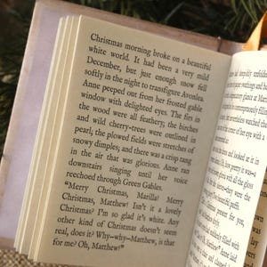 May include: An open miniature book with cream-colored pages featuring black printed text from Anne of Green Gables. The book has a light-colored cover and a thin gold ribbon bookmark attached to the spine, displaying a clear view of the narrative.