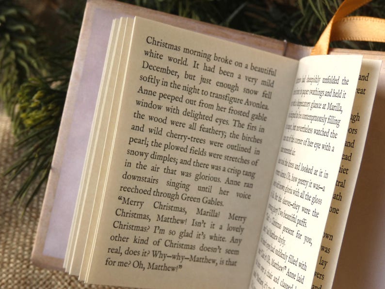 May include: An open miniature book with cream-colored pages featuring black printed text from Anne of Green Gables. The book has a light-colored cover and a thin gold ribbon bookmark attached to the spine, displaying a clear view of the narrative.
