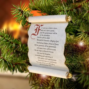 May include: A hanging scroll ornament featuring a parchment-style paper with a decorative red initial letter and black printed text that reads For unto us a child is born, unto us a son is given, and the government will be on his shoulders.