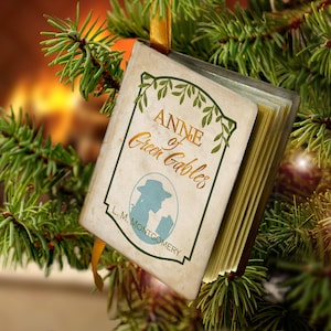 May include: A miniature book ornament titled Anne of Green Gables by L. M. Montgomery, featuring a cream-colored cover with green leaf illustrations, a blue silhouette of a person in a hat, and a gold ribbon for hanging on a tree.
