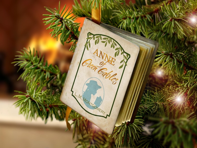 May include: A miniature book ornament titled Anne of Green Gables by L. M. Montgomery, featuring a cream-colored cover with green leaf illustrations, a blue silhouette of a person in a hat, and a gold ribbon for hanging on a tree.