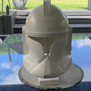 Puede incluir: Un casco de plástico gris claro, que recuerda a un casco de soldado clon, se encuentra sobre un salvamanteles redondo y tejido. El casco tiene una parte superior redondeada con una cresta vertical y una protección facial detallada.