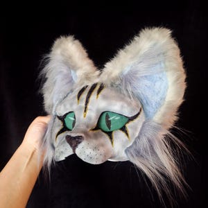 May include: A handmade cat mask with a white and gray fur design. The mask features green mesh eyes, black and gold accents, and a black nose. The ears are lined with blue fabric.