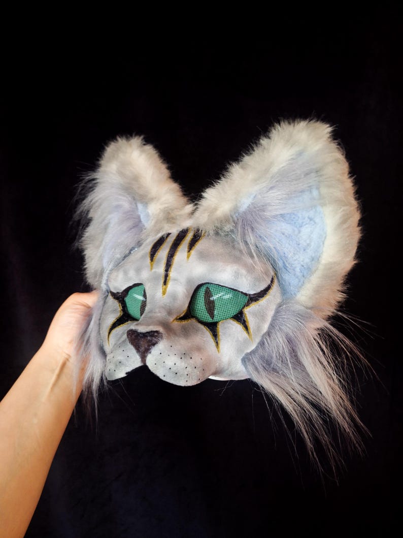 May include: A handmade cat mask with a white and gray fur design. The mask features green mesh eyes, black and gold accents, and a black nose. The ears are lined with blue fabric.