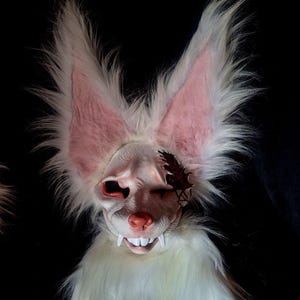 Op de afbeelding: Een wit imitatiebont masker met grote, puntige oren met roze voering, een gebeeldhouwde snuit met een roze neus en opvallende witte tanden. Het masker bevat een donker, grillig, vleugelachtig decoratief element over de linker oogkas.