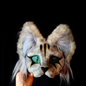 May include: A gray cat mask with green eyes and black markings. The mask has fluffy ears and is held against a black background. The cat's face has yellow stripes and black star-shaped markings around the eyes.