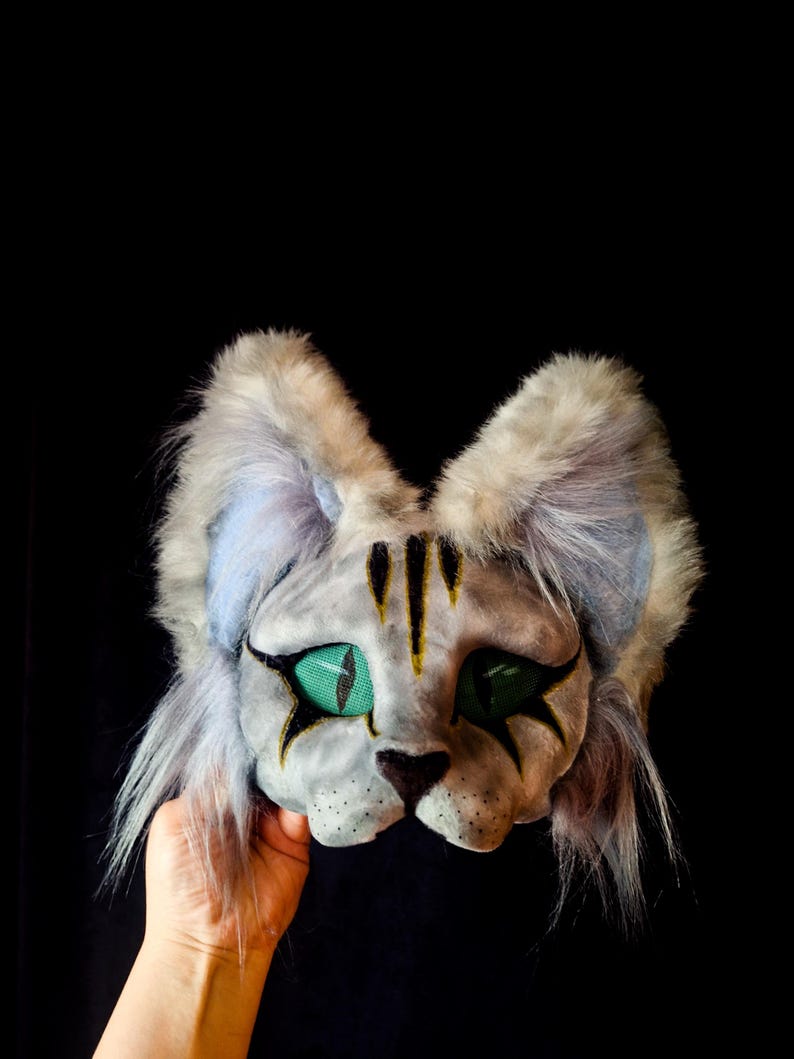 May include: A gray cat mask with green eyes and black markings. The mask has fluffy ears and is held against a black background. The cat's face has yellow stripes and black star-shaped markings around the eyes.