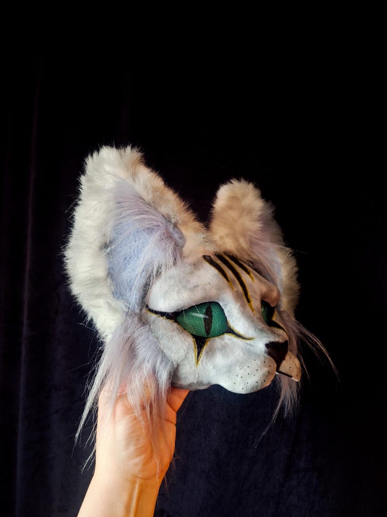 May include: A white and gray cat mask with green eyes and black markings. The mask has fluffy fur and long whiskers. The mask is held up by a hand against a black background.