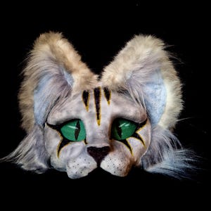 May include: A cat mask with a white and gray face, green mesh eyes, and black and gold accents. The mask has fluffy gray and blue ears and fur around the face. The cat's nose is black.