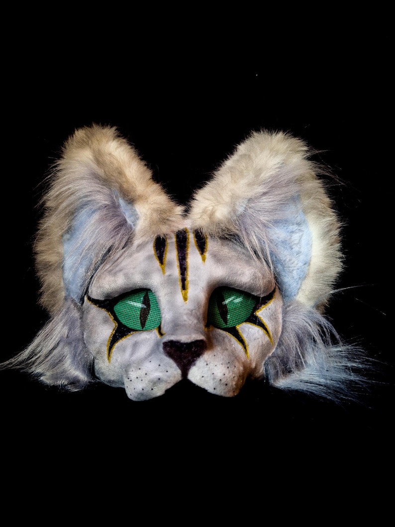 May include: A cat mask with a white and gray face, green mesh eyes, and black and gold accents. The mask has fluffy gray and blue ears and fur around the face. The cat's nose is black.
