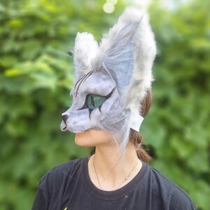 May include: A gray and white cat mask with large, fluffy ears and green eyes. The mask is worn over the face, with a strap around the back of the head. The mask has a detailed, painted design.