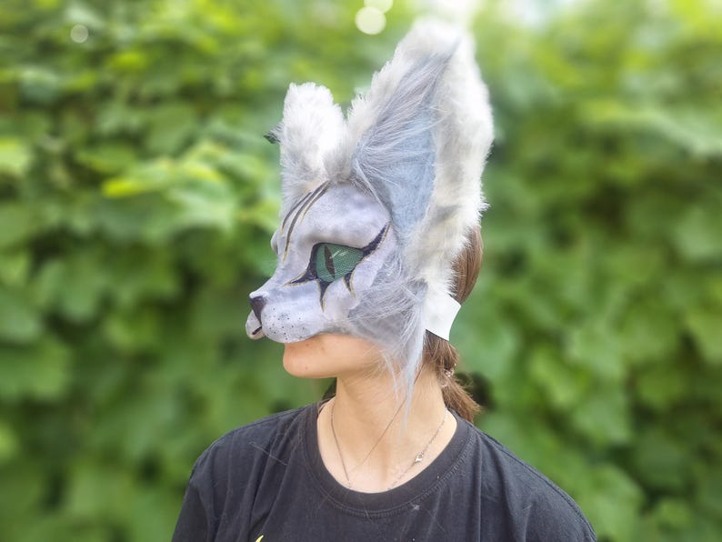 May include: A gray and white cat mask with large, fluffy ears and green eyes. The mask is worn over the face, with a strap around the back of the head. The mask has a detailed, painted design.