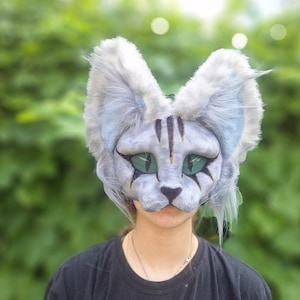 May include: A detailed cat mask with a gray and white color scheme, featuring large, expressive green eyes and black markings. The mask has large, fluffy ears and is designed to cover the face. The background is a blurred green foliage.