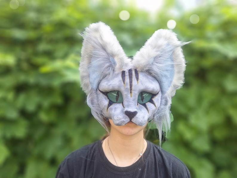 May include: A detailed cat mask with a gray and white color scheme, featuring large, expressive green eyes and black markings. The mask has large, fluffy ears and is designed to cover the face. The background is a blurred green foliage.
