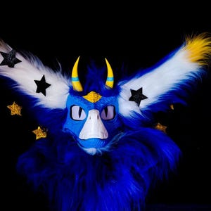 Può includere: Una maschera di creatura pelosa blu, bianca e gialla con grandi ali piumate. La maschera ha corna gialle, dettagli di stelle nere e stelle glitterate dorate. Lo sfondo è nero.