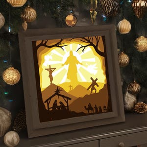 May include: A framed, layered paper art piece depicting a religious scene. The artwork features a silhouette of a figure with outstretched arms, a cross, and other biblical elements. The color palette includes shades of brown and yellow, with a wooden frame.