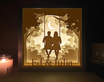 May include: A framed, layered paper art piece featuring a silhouette of a couple on a swing under a crescent moon. The artwork is illuminated from within, creating a warm, inviting glow. The names "Ed & Julia" are cut into the bottom.