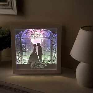 May include: A white framed shadow box featuring a silhouette of a couple on their wedding day. The scene includes a moon, stars, and floral accents. The date "09-29-2012" is visible. The box is illuminated with soft, colorful lighting.