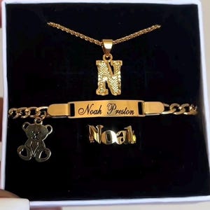 May include: A gold-coloured jewellery set presented in a black box. The set includes a necklace with the letter "N" pendant, a bracelet engraved with "Noah Preston", and a ring spelling "Noal". A teddy bear charm hangs from the bracelet.