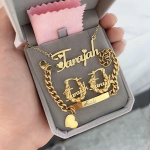 May include: A gold-colored jewelry set including a necklace, bracelet, and earrings, all with the name "Tarajah" and a heart design. The set is presented in a gray box with a pink cloth.