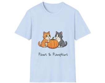 Könnte beinhalten: Hellblaues T-Shirt mit einer Cartoon-Illustration von zwei Katzen und einem Kürbis. Eine Katze ist orange und weiß, die andere grau und weiß. Der Text "Paws & Pumpkins" ist darunter aufgedruckt.