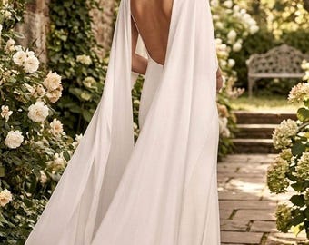 Puede incluir: Un vestido blanco largo hasta el suelo con una espalda profundamente abierta y mangas largas y fluidas estilo capa que caen desde los hombros hasta el suelo. El vestido está confeccionado con una tela blanca lisa y sólida con una silueta limpia.